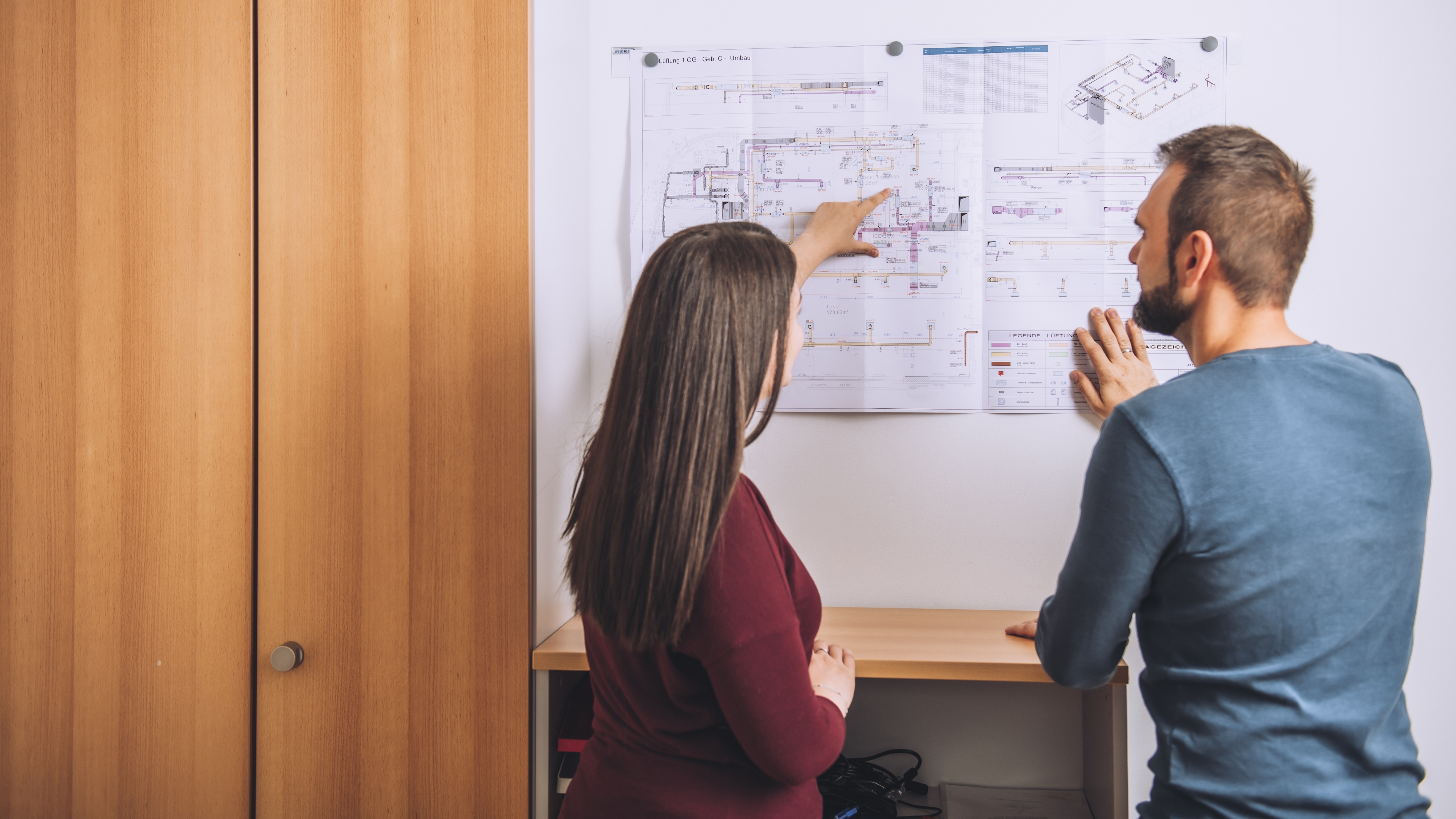 Ausbildung Technischer Systemplaner: Vom leeren Bildschirm zum fertigen Gebäudeplan Junge Frau zeigt etwas auf einem Plan, der an der Wand hängt, einem Kollegen
